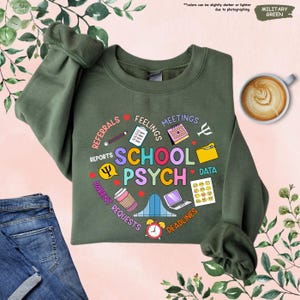 May include: A green sweatshirt featuring a graphic design of a speech bubble that reads "School Psych" surrounded by icons and text related to the work of a school psychologist. The text includes "Referrals", "Feelings", "Meetings", "Reports", "Parent Requests", "Data", and "Deadlines".