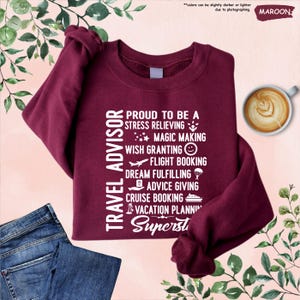 May include: A maroon sweatshirt with white text that reads "Travel Advisor" and lists the duties of a travel advisor, including "stress relieving", "magic making", "wish granting", "flight booking", "dream fulfilling", "advice giving", "cruise booking", and "vacation planning". The text ends with the word "Superst".