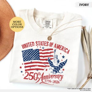 Op de afbeelding: Ivoor kleurig t-shirt met een verweerd Amerikaans vlagontwerp met de tekst "UNITED STATES OF AMERICA" en "250th Anniversary 1776-2026." De shirt bevat een afbeelding van het Vrijheidsbeeld en een adelaar. Een gouden ketting ligt op het shirt.