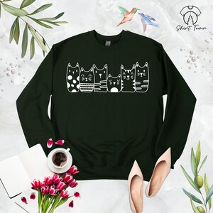 Puede incluir: Una sudadera verde oscuro con un gr&aacute;fico blanco de cinco gatos en fila. Los gatos son todos de dise&ntilde;os diferentes, uno lleva lunares, uno rayas, uno con un dise&ntilde;o simple y dos con rayas.