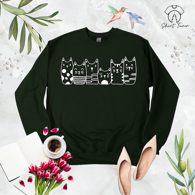 Puede incluir: Una sudadera verde oscuro con un gr&aacute;fico blanco de cinco gatos en fila. Los gatos son todos de dise&ntilde;os diferentes, uno lleva lunares, uno rayas, uno con un dise&ntilde;o simple y dos con rayas.