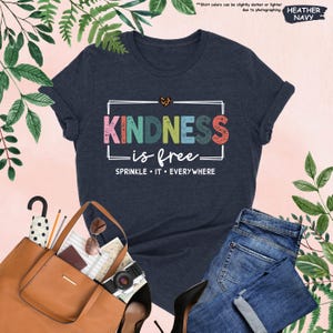May include: A heather navy blue t-shirt with a white rectangle around the text "KINDNESS is free" in rainbow colors with a small heart above the text. The text "SPRINKLE IT EVERYWHERE" is below the rectangle.