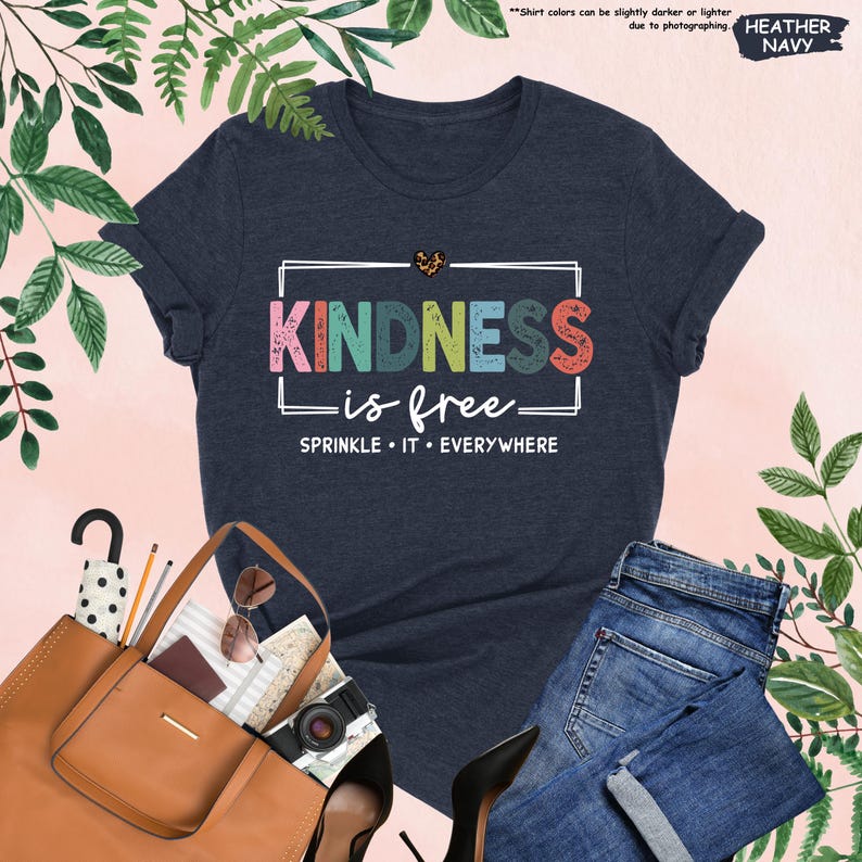 May include: A heather navy blue t-shirt with a white rectangle around the text "KINDNESS is free" in rainbow colors with a small heart above the text. The text "SPRINKLE IT EVERYWHERE" is below the rectangle.