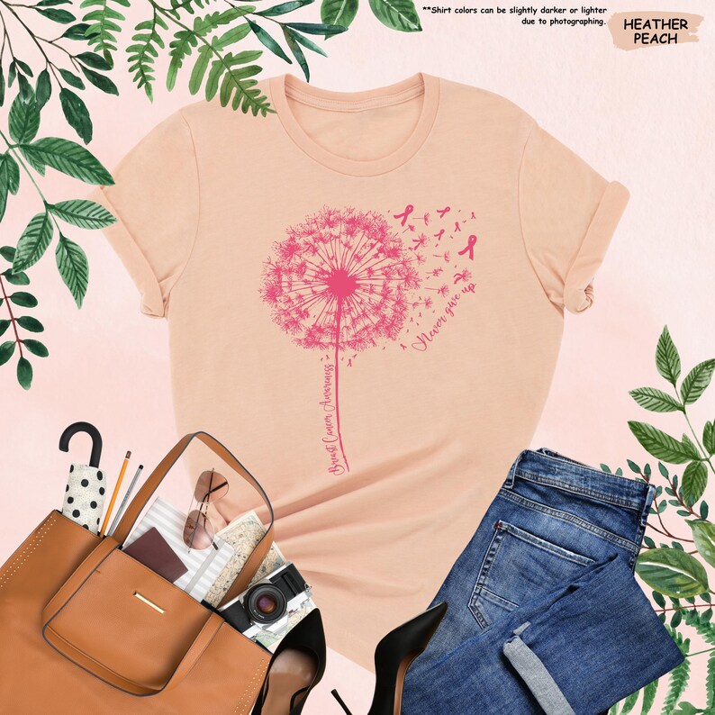 Puede incluir: Camiseta color melocot&oacute;n con un dise&ntilde;o de diente de le&oacute;n rosa, lazos rosas y las palabras "Breast Cancer Awareness" y "Never give up." La camiseta se combina con vaqueros azules, un bolso marr&oacute;n y tacones negros.