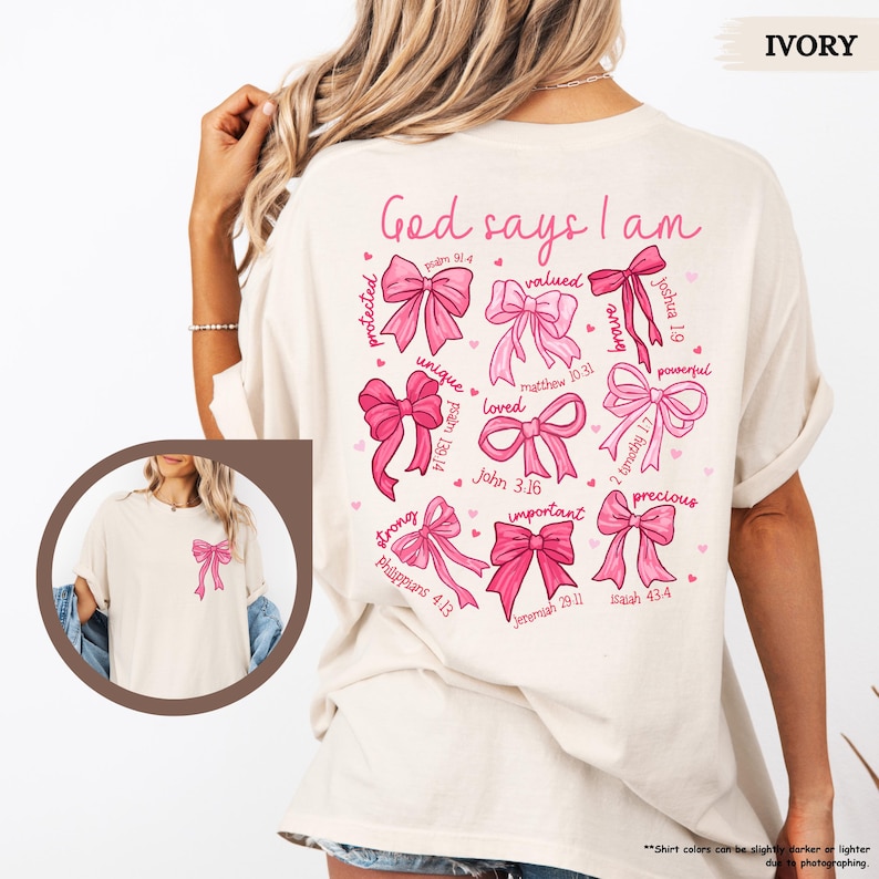 K&ouml;nnte beinhalten: Ein T-Shirt in Elfenbein mit einem rosafarbenen Grafikdesign aus Schleifen und Bibelversen. Der Text lautet "Gott sagt, ich bin" und enth&auml;lt Verse wie "Psalm 91:4", "Matth&auml;us 10:31" und "Johannes 3:16".