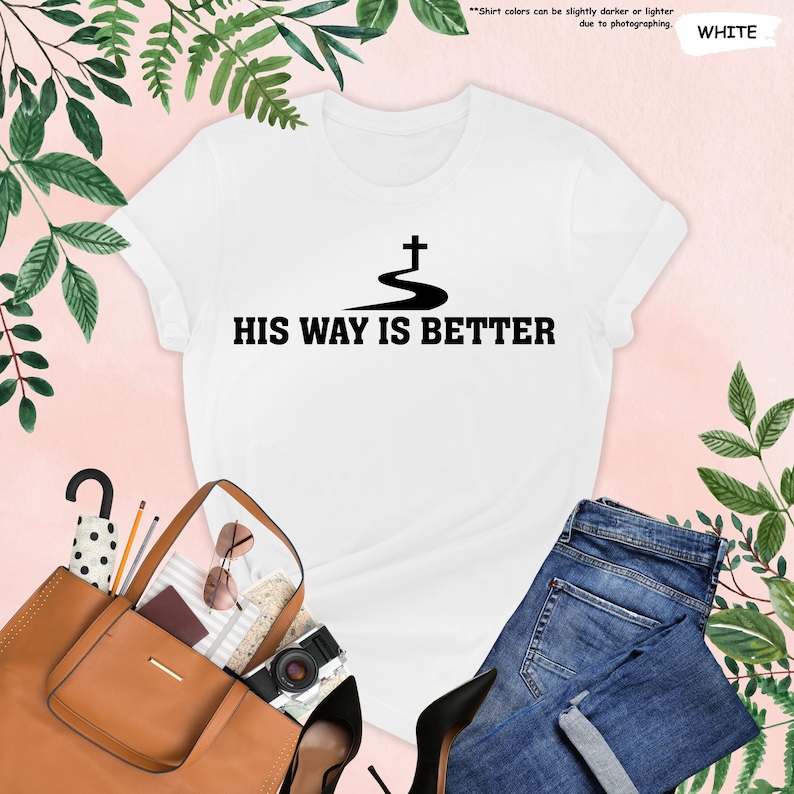 May include: White t-shirt with the text "HIS WAY IS BETTER" in black, featuring a cross design. The shirt is paired with blue jeans, a brown purse, black heels, and accessories. The background is a soft pink with green foliage.