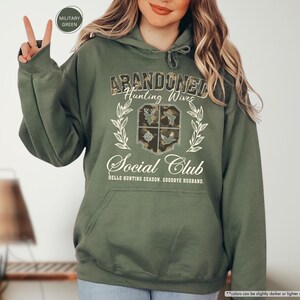 May include: Military green hoodie with the text "ABANDONED Hunting Wives Social Club" in a decorative font. The design includes a crest with hunting-themed imagery and the phrase "HELLO HUNTING SEASON, GOODBYE HUSBAND."