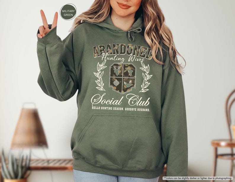 May include: Military green hoodie with the text "ABANDONED Hunting Wives Social Club" in a decorative font. The design includes a crest with hunting-themed imagery and the phrase "HELLO HUNTING SEASON, GOODBYE HUSBAND."