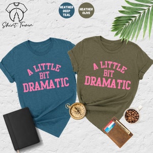 May include: Two t-shirts with the text "A Little Bit Dramatic" in pink lettering on a teal and olive green background.
