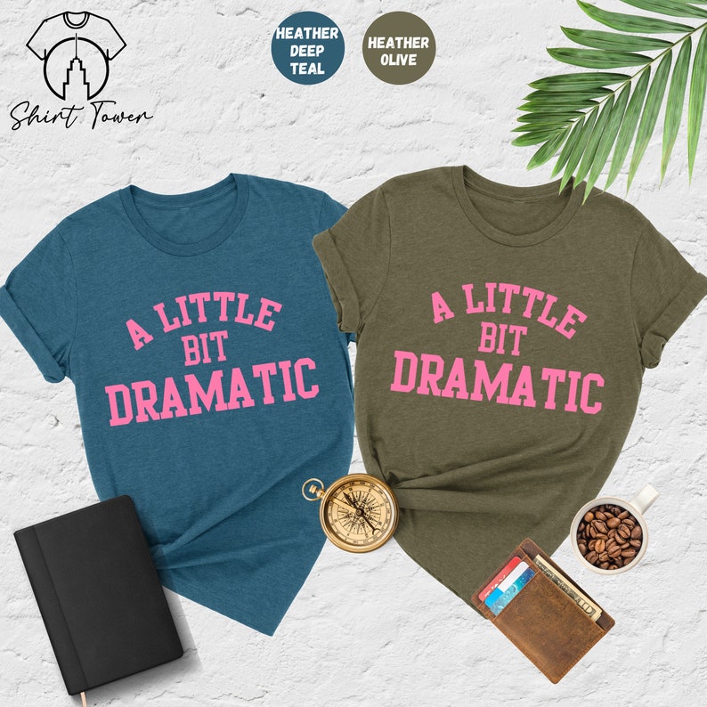 May include: Two t-shirts with the text "A Little Bit Dramatic" in pink lettering on a teal and olive green background.
