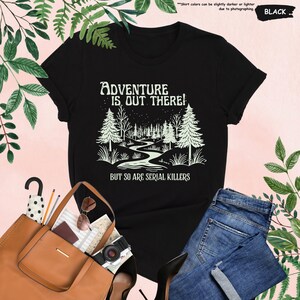 Op de afbeelding: Zwart t-shirt met de tekst "ADVENTURE IS OUT THERE! BUT SO ARE SERIAL KILLERS" en een bosafbeelding. Het shirt wordt gecombineerd met een blauwe spijkerbroek en een bruine tote bag.