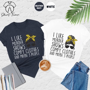May include: Two t-shirts with a graphic of a woman with a messy bun and sunglasses. The text on the shirts reads "I like murder shows comfy clothes and maybe 3 people". The shirts are available in heather navy and white.