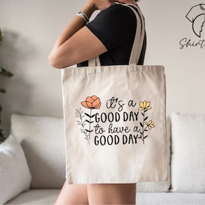 May include: A white canvas tote bag with the text "it's a good day to have a good day" in black lettering. The text is surrounded by small orange and yellow flowers.