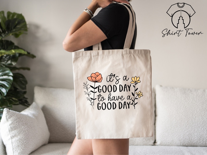 May include: A white canvas tote bag with the text "it's a good day to have a good day" in black lettering. The text is surrounded by small orange and yellow flowers.