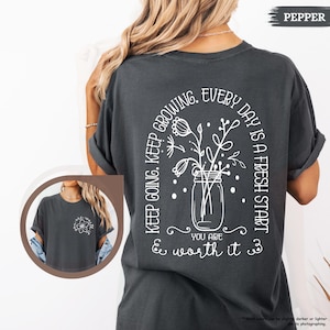 May include: A dark grey t-shirt with a white graphic of a jar with flowers and the text "Keep going. Keep growing. Every day is a fresh start. You are & worth it."
