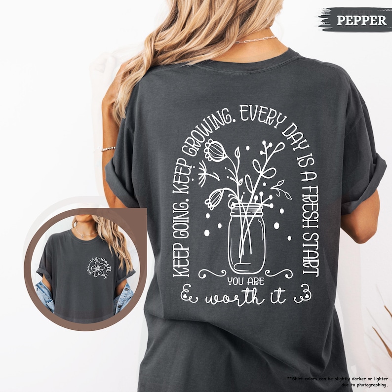 May include: A dark grey t-shirt with a white graphic of a jar with flowers and the text "Keep going. Keep growing. Every day is a fresh start. You are & worth it."