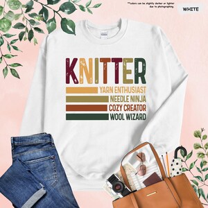 May include: White sweatshirt with the word "KNITTER" in brown, red, and yellow letters. Below the word are four lines of text in a vintage style font: "YARN ENTHUSIAST", "NEEDLE NINJA", "COZY CREATOR", and "WOOL WIZARD".