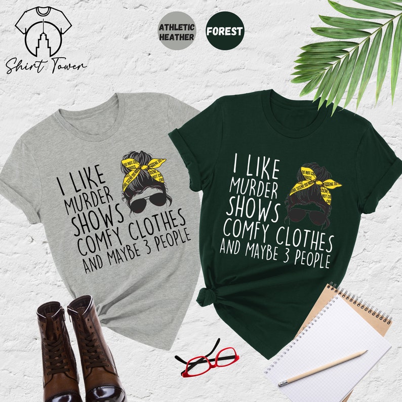 May include: Two t-shirts with a graphic of a woman with a messy bun and sunglasses. The text on the shirts reads "I like murder shows, comfy clothes and maybe 3 people". The shirts are in athletic heather grey and forest green.