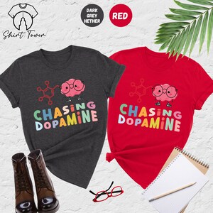 Pu&ograve; includere: Due t-shirt con un cervello di cartone animato che indossa gli occhiali e il testo "Chasing Dopamine" in colori arcobaleno. Le t-shirt sono in grigio melange scuro e rosso.