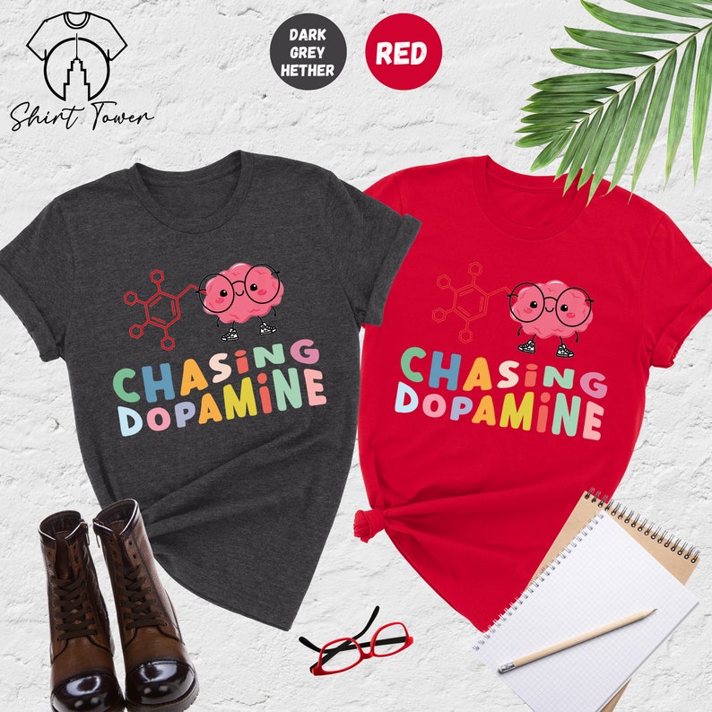 Pu&ograve; includere: Due t-shirt con un cervello di cartone animato che indossa gli occhiali e il testo "Chasing Dopamine" in colori arcobaleno. Le t-shirt sono in grigio melange scuro e rosso.