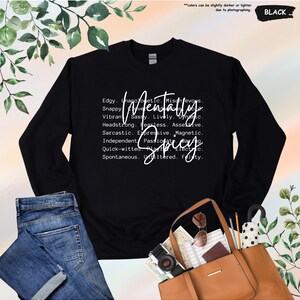 May include: Black sweatshirt with the words "Mentally Spicy" in white script. The design includes a list of adjectives such as "edgy," "sassy," and "fearless." The sweatshirt is paired with blue jeans and a brown tote bag.