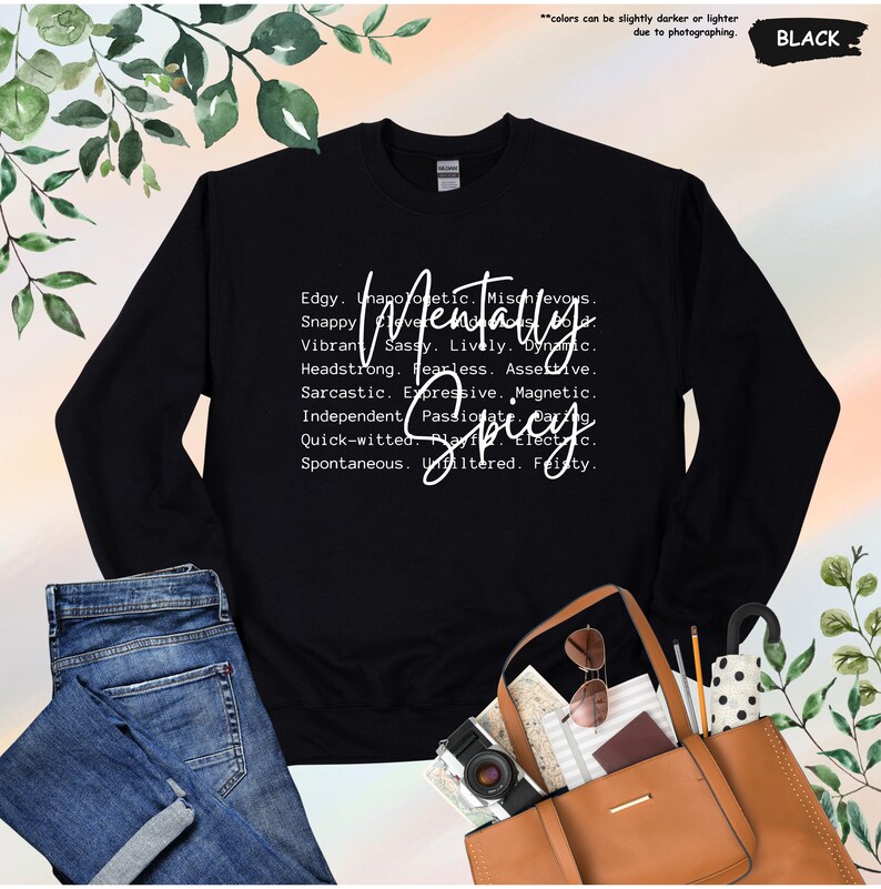 May include: Black sweatshirt with the words "Mentally Spicy" in white script. The design includes a list of adjectives such as "edgy," "sassy," and "fearless." The sweatshirt is paired with blue jeans and a brown tote bag.