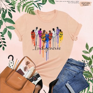 May include: A peach colored t-shirt with a graphic of a group of people with feathers in their hair. The feathers are dripping with colorful paint and the word "Indigenous" is written below the graphic.
