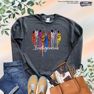 May include: Dark heather grey sweatshirt with a colorful design of people holding feathers and the word "Indigenous" in a dripping paint style.