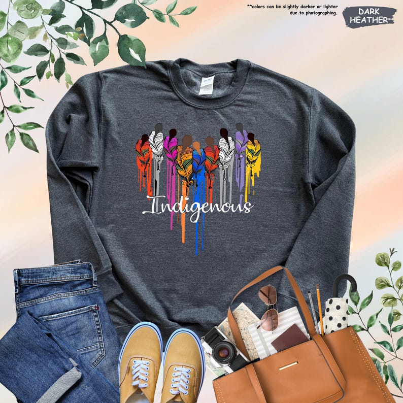 May include: Dark heather grey sweatshirt with a colorful design of people holding feathers and the word "Indigenous" in a dripping paint style.
