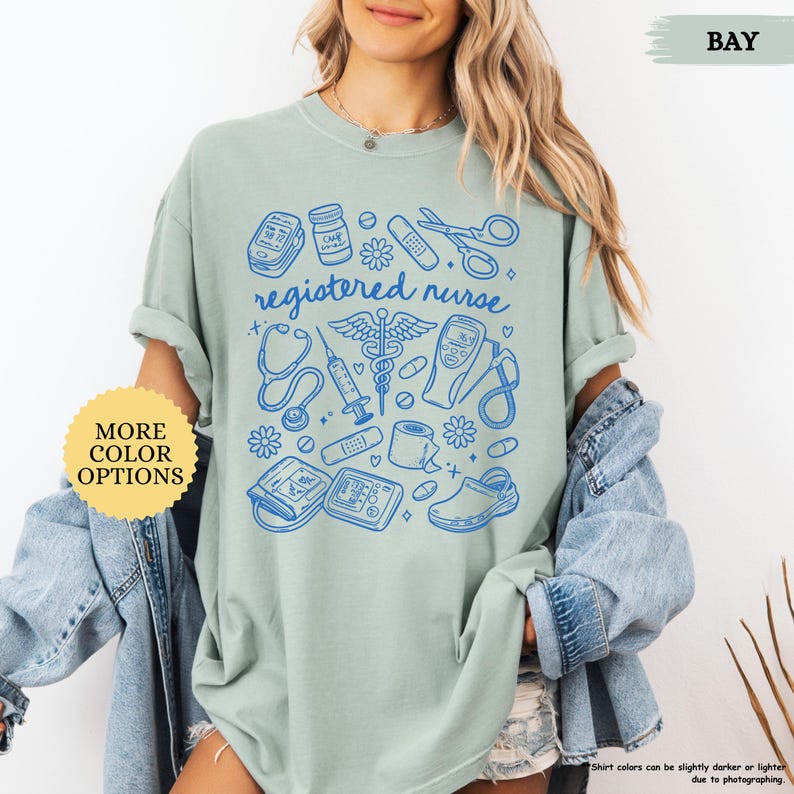 May include: A light green t-shirt with a blue graphic design featuring medical tools and the words "registered nurse." The design includes a stethoscope, syringe, and other medical symbols. The shirt has rolled-up sleeves and a relaxed fit.
