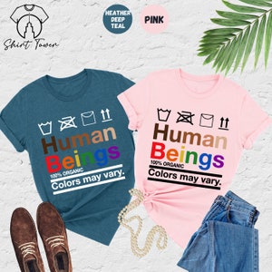 May include: Two t-shirts with the text "Human Beings 100% Organic Colors may vary." The text is in rainbow colors and is surrounded by laundry care symbols. One shirt is heather deep teal and the other is pink.