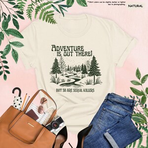 Op de afbeelding: Cr&egrave;mekleurig T-shirt met de tekst "ADVENTURE IS OUT THERE! BUT SO ARE SERIAL KILLERS" en een bosafbeelding. Het shirt wordt gecombineerd met een blauwe jeans, een bruine handtas en zwarte hakken.