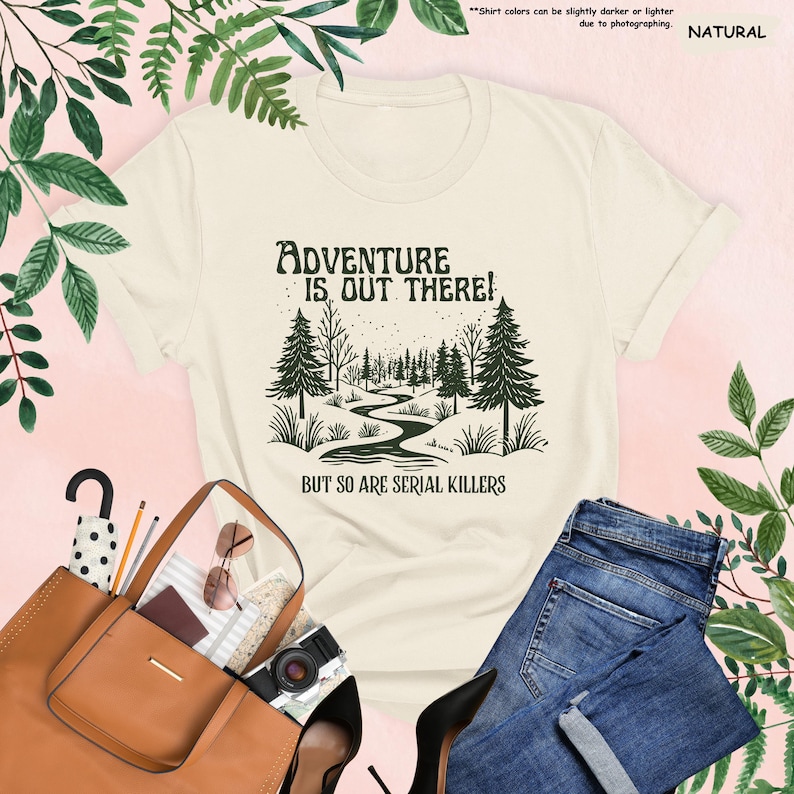 Op de afbeelding: Cr&egrave;mekleurig T-shirt met de tekst "ADVENTURE IS OUT THERE! BUT SO ARE SERIAL KILLERS" en een bosafbeelding. Het shirt wordt gecombineerd met een blauwe jeans, een bruine handtas en zwarte hakken.