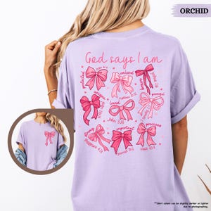 K&ouml;nnte beinhalten: Ein helles lila T-Shirt mit dem Text "God says I am" auf der R&uuml;ckseite. Der Text ist umgeben von rosa Schleifen mit Bibelversen und W&ouml;rtern wie "gesch&uuml;tzt", "gesch&auml;tzt", "einzigartig", "geliebt", "stark", "wichtig", "m&auml;chtig" und "kostbar".