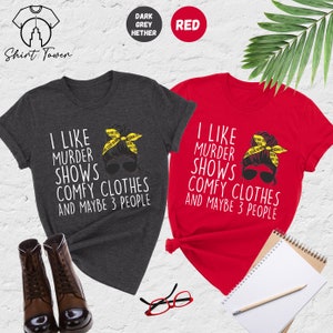 May include: Two t-shirts with a graphic of a woman with a messy bun wearing sunglasses and a yellow crime scene tape headband. The text on the shirts reads "I like murder shows comfy clothes and maybe 3 people". One shirt is dark heather grey and the other is red.