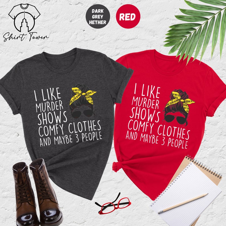 May include: Two t-shirts with a graphic of a woman with a messy bun wearing sunglasses and a yellow crime scene tape headband. The text on the shirts reads "I like murder shows comfy clothes and maybe 3 people". One shirt is dark heather grey and the other is red.