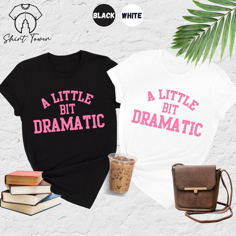 May include: Two t-shirts, one black and one white, with the text "A Little Bit Dramatic" in pink letters. The shirts are laid out on a surface with a cup of iced coffee and a brown purse.