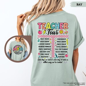 May include: A light green t-shirt with a colorful graphic that says "Teacher Tour" and lists tasks teachers do, such as "Multi Tasking", "Book Readin", "Lesson Plannin", "Behavior Managin", "Pencil Sharpenin", "Cafeteria Goin", "Song Singin", "Negotiatin", "Conferencing", "Paper Gradin", "Snack Dealin", "Coffee Drinkin", "Field Trip Plannin", "Recess Monitorin", "Evaluatin", and "Nurturin". The graphic also includes a red apple, a yellow lightning bolt, and a yellow smiley face.