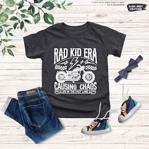 May include: A dark grey heather t-shirt with a white graphic of a motorcycle and the text "Rad Kid Era Causing Chaos Life in the Fast Lane".