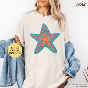 May include: Ivory-colored t-shirt featuring a large, detailed starfish design. The starfish is teal with pink floral accents and a central orange starfish. The shirt has the text "MORE COLOR OPTIONS" on a yellow circle.