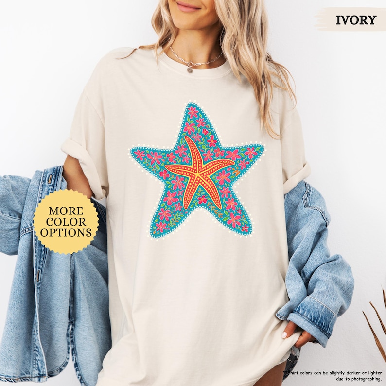 May include: Ivory-colored t-shirt featuring a large, detailed starfish design. The starfish is teal with pink floral accents and a central orange starfish. The shirt has the text "MORE COLOR OPTIONS" on a yellow circle.