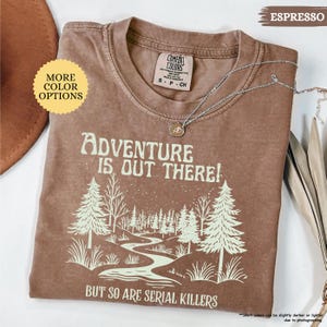 May include: Espresso-colored t-shirt with the text "ADVENTURE IS OUT THERE! BUT SO ARE SERIAL KILLERS" and a forest graphic. The shirt has a crew neck and short sleeves. A silver necklace with a pendant is draped across the shirt.