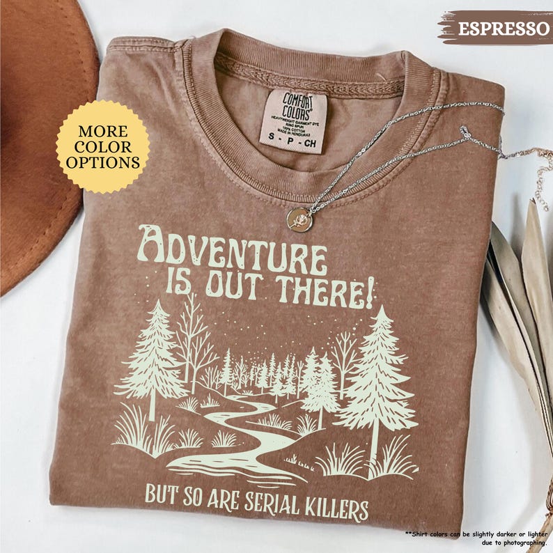 May include: Espresso-colored t-shirt with the text "ADVENTURE IS OUT THERE! BUT SO ARE SERIAL KILLERS" and a forest graphic. The shirt has a crew neck and short sleeves. A silver necklace with a pendant is draped across the shirt.