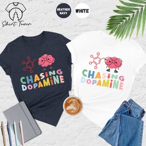 Pu&ograve; includere: Due t-shirt con una grafica colorata di un cervello con gli occhiali e una struttura chimica. Il testo "Chasing Dopamine" &egrave; scritto in colori arcobaleno su entrambe le magliette. Una maglietta &egrave; blu navy melange e l'altra &egrave; bianca.