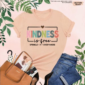 May include: A peach colored t-shirt with the text "KINDNESS is free" in rainbow colors and a heart with leopard print. The text "SPRINKLE IT EVERYWHERE" is below the main text.