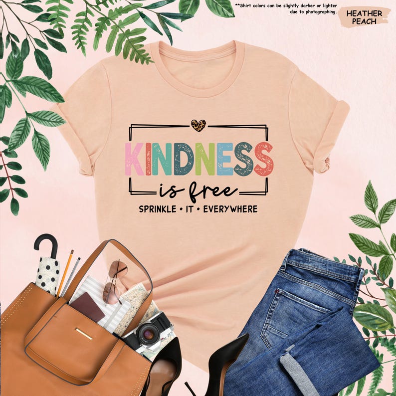 May include: A peach colored t-shirt with the text "KINDNESS is free" in rainbow colors and a heart with leopard print. The text "SPRINKLE IT EVERYWHERE" is below the main text.