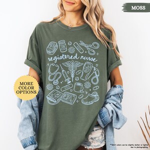 May include: Moss green t-shirt with a light blue graphic of medical tools and the words "registered nurse." The design includes a stethoscope, syringe, and other medical symbols. A yellow circle with the words "MORE COLOR OPTIONS" is on the left.