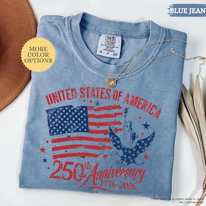 Op de afbeelding: Lichtblauw T-shirt met een patriottisch ontwerp ter viering van de 250e verjaardag van de Verenigde Staten. De afbeelding bevat de Amerikaanse vlag, het Vrijheidsbeeld en een adelaar. De tekst luidt "UNITED STATES OF AMERICA 250th Anniversary 1776-2026."