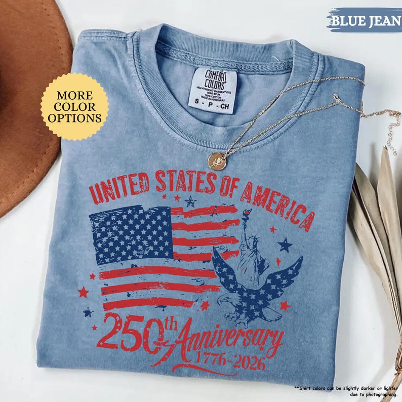 Op de afbeelding: Lichtblauw T-shirt met een patriottisch ontwerp ter viering van de 250e verjaardag van de Verenigde Staten. De afbeelding bevat de Amerikaanse vlag, het Vrijheidsbeeld en een adelaar. De tekst luidt "UNITED STATES OF AMERICA 250th Anniversary 1776-2026."