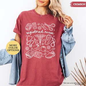 May include: Crimson-colored t-shirt with the words "registered nurse" and medical-related graphics in white. The design includes a stethoscope, syringe, and other medical tools. The shirt has a relaxed fit and rolled-up sleeves.